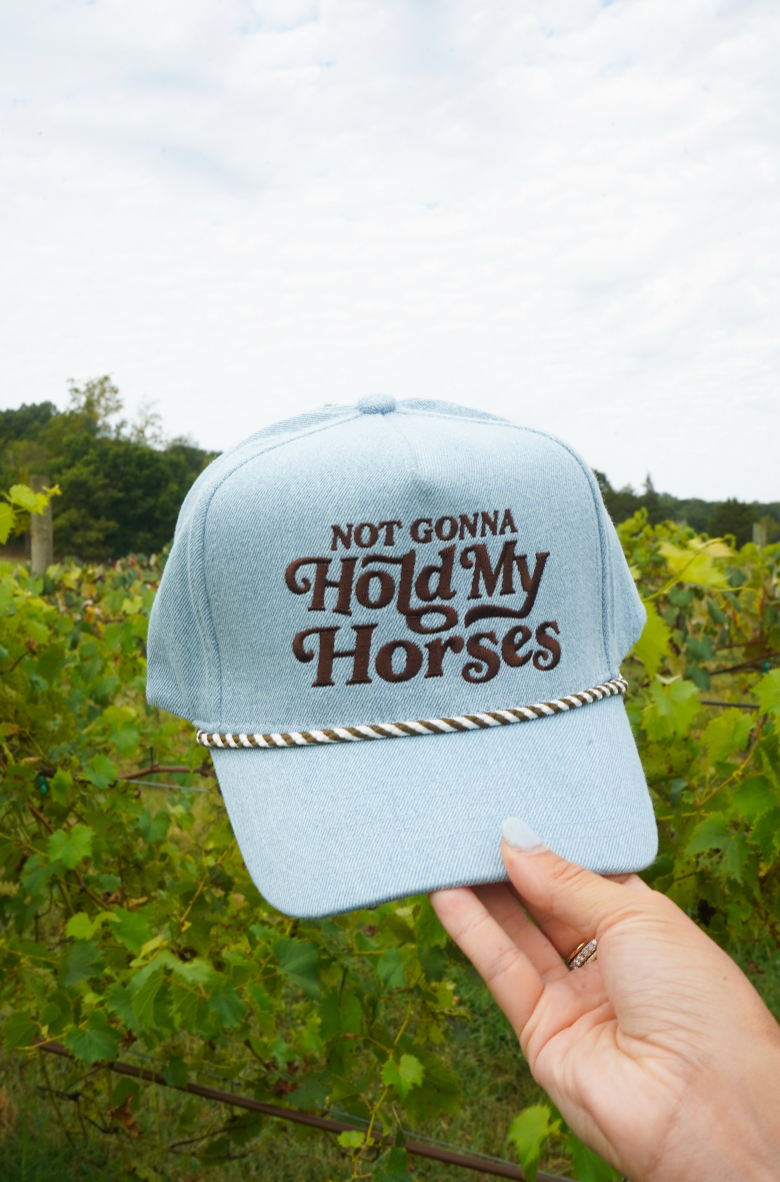 A light blue denim baseball cap held by a hand in an outdoor vineyard setting. The cap features brown embroidered text reading &